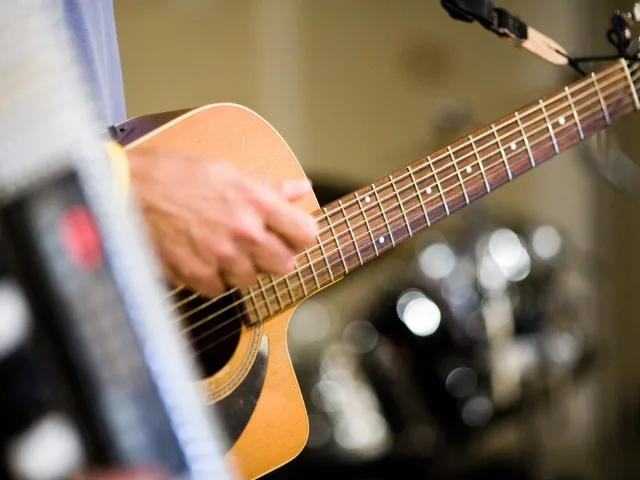 Close-up of a person playing an acoustic guitar. The focus is on the hand strumming the strings and the body of the guitar. The background is blurred, highlighting the instrument and the musician's hand. Reflection of lights creates a bokeh effect in the background.