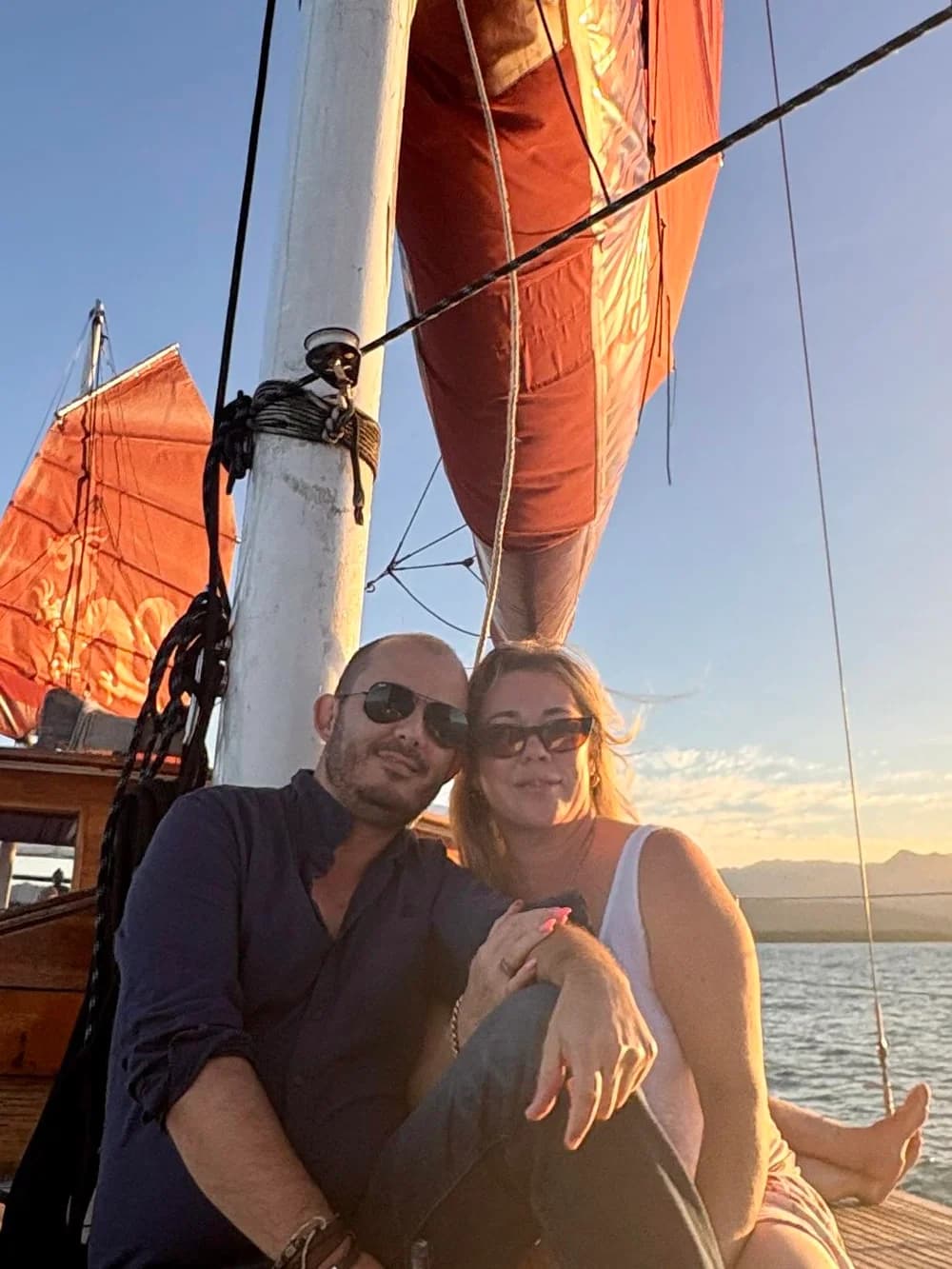Two people aboard a sailing boat beneath orange sails in the evening sunlight.